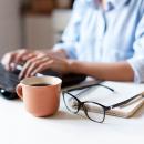 一位女士坐在笔记本电脑前，旁边放着一杯咖啡、笔记本电脑和一副眼镜，正在打字